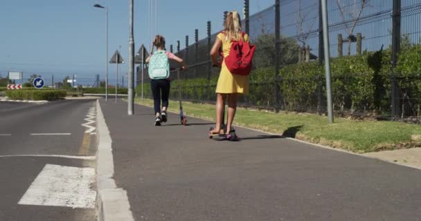 2人の白人の女の子が学校に行く途中で学校の袋を持って舗装されたスクーターを押して ゆっくりと動きます 小学生 道路安全 — ストック動画
