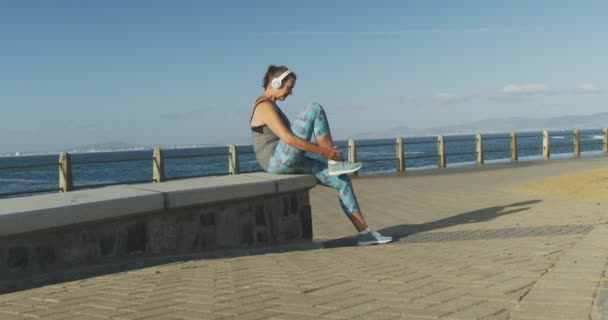 スポーツウェアを着て海の遊歩道で働くシニア白人女性は ゆっくりとした動きでヘッドフォンで靴ひもを結ぶ休憩を取ります 退職健康的なライフスタイル活動 — ストック動画