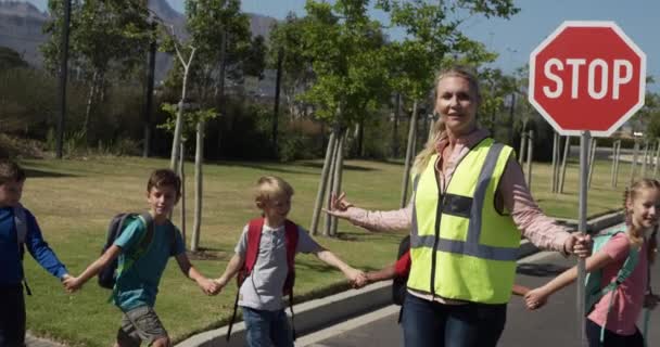 身穿高光背心的白人妇女 手持停车标志 站在人行横道上 多族裔儿童走过马路 行动迟缓 小学生 道路安全 — 图库视频影像