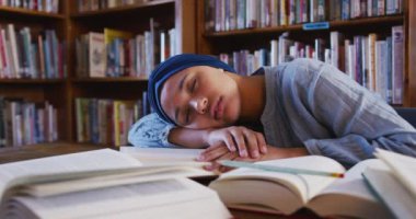 Mavi başörtüsü takan Asyalı bir kız öğrenci açık kitaplarla dolu bir masada oturuyor, kollarına uzanıyor ve uyuyor, ağır çekimde günlük kıyafetler giyiyor. Öğrenci çok çalışmaktan yoruldu..