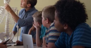 Kafkas erkek öğretmen, çok ırklı bir grup çocuğa biyoloji dersi sırasında yavaş çekimde insan kemiklerini öğretmek için insan iskeleti modelini kullanıyor. İlkokulda eğitim.