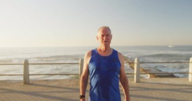 Deniz kenarındaki gezinti güvertesinde spor kıyafetleri giymiş, mola verip ağır çekimde gülümseyen yaşlı bir beyaz adamın portresi. Emeklilik sağlıklı yaşam tarzı aktivitesi.