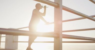 Kıdemli beyaz adam deniz kenarında spor kıyafetleri giyerek gezinti yapıyor, mola veriyor ve ağır çekimde su içiyor. Emeklilik sağlıklı yaşam tarzı aktivitesi.