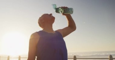 Kıdemli beyaz adam, deniz kenarında spor kıyafetleri giyerek gezinti yapıyor, mola veriyor ve yavaş çekimde serinliyor. Emeklilik sağlıklı yaşam tarzı aktivitesi.