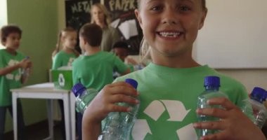 Üzerinde geri dönüşüm logosu olan beyaz bir kızın portresi, elinde plastik şişeler, arka planda sınıf arkadaşları, yavaş çekim, bir ilkokulda eğitim....
