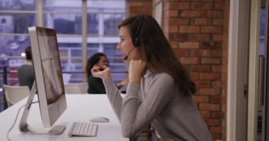 Modern bir ofiste çalışan, telefon kulaklığı takan, bilgisayar kullanan, arka planda çalışan beyaz profesyonel iş kadını. İş yaratıcılığı teknolojisi.