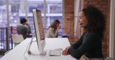 Modern bir ofiste çalışan, telefon kulaklığı takan, bilgisayar kullanan ve arka planda birlikte çalışan melez bir iş kadını. İş yaratıcılığı teknolojisi.