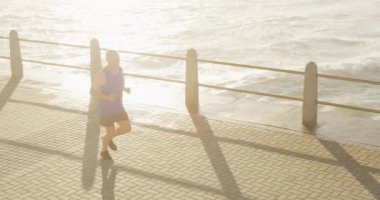 Kıdemli beyaz adam deniz kenarındaki gezinti güvertesinde spor kıyafetleriyle çalışıyor, güneşli bir günde ağır çekimde koşuyor. Emeklilik sağlıklı yaşam tarzı aktivitesi.