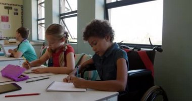 Afrika kökenli Amerikalı ve Kafkasyalı kızlar ders sırasında sınıfta oturup not defterlerine yazıyorlar, biri ağır çekimde tekerlekli sandalyede oturuyor. İlkokulda eğitim.