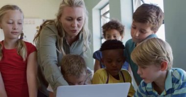 Çok ırklı çocuklardan oluşan bir grup ve ders sırasında sınıfta ağır çekimde dizüstü bilgisayar kullanarak oturan beyaz kadın öğretmenleri. İlkokulda eğitim.