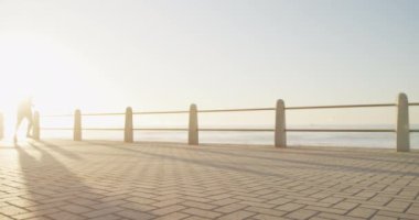 Kıdemli beyaz adam deniz kenarındaki gezinti güvertesinde spor kıyafetleriyle çalışıyor, güneşli bir günde ağır çekimde koşuyor. Emeklilik sağlıklı yaşam tarzı aktivitesi.