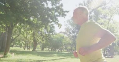 Kıdemli beyaz adam parkta spor kıyafetleriyle çalışıyor, güneşli bir günde ağır çekimde koşuyor. Emeklilik sağlıklı yaşam tarzı aktivitesi.