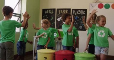 Beyaz geri dönüşüm logosu olan yeşil tişörtler giyen, yavaş çekimde beşlik çakan ve gülümseyen çok ırklı çocuklar. Bir ilkokulda eğitim.