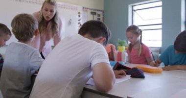 Ders sırasında sınıfta oturan beyaz bir çocuğun portresi, arka planda sınıf arkadaşları ve öğretmeniyle birlikte kameraya bakıyor ve gülümsüyor. İlkokulda eğitim.