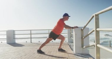 Deniz kenarındaki gezinti güvertesinde spor giysileri giymiş güneşli bir günde ağır çekimde uzanan kıdemli beyaz adam. Emeklilik sağlıklı yaşam tarzı aktivitesi.