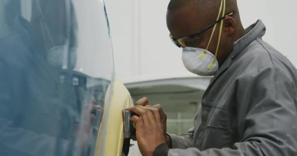 アフリカ系アメリカ人の男性の車の整備士は 町のワークショップで働いて 顔のマスクを着用し スローモーションで車の側面に研磨機を使用して — ストック動画