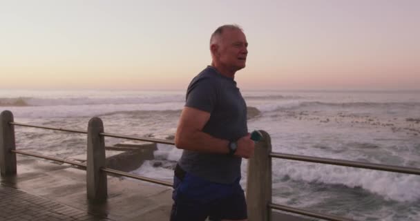 シニアフィットゆっくりとした動きで晴れた日に実行され スポーツ服を着て海で遊歩道に取り組んで白人男性 退職健康的なライフスタイル活動 — ストック動画