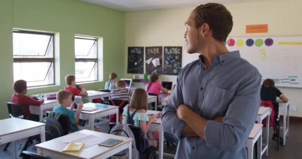 両腕を組んで教室に立つ白人男性教師は カメラを見て笑顔で 生徒たちと机の後ろに座ってゆっくりとした動きをしていた 小学校での教育 — ストック動画