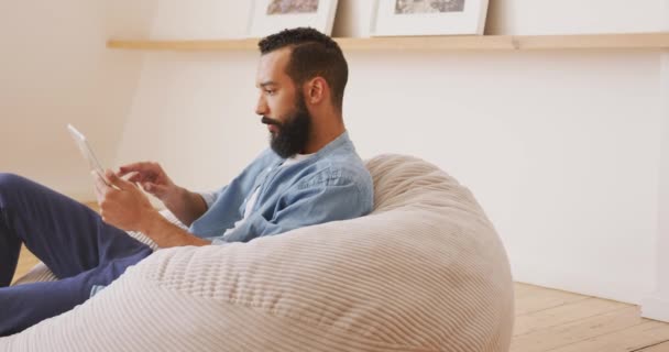 混血男子在家里休息 坐在豆袋上 使用数码平板电脑 — 图库视频影像