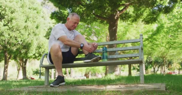 シニアフィット白人男性は公園でスポーツ服を着て作業し ベンチに座ってゆっくりとした動きで自分の靴を結びます 退職健康的なライフスタイル活動 — ストック動画