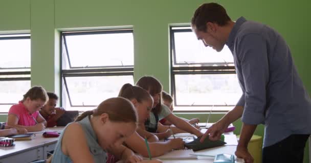 机の前に立つ白人男性の学校教師は 生徒たちがゆっくりと机の上に座っているのを助ける 小学校での教育 — ストック動画