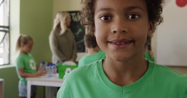 非洲裔美国女孩的画像 身穿绿色T恤 带有回收标识 看着相机 背景是她的同学和老师 动作缓慢 小学的教育 — 图库视频影像