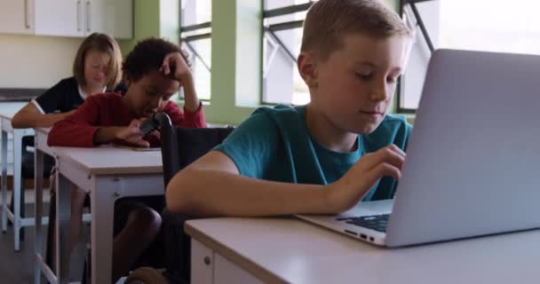 教室の机に座っている白人の少年は ラップトップコンピュータを使用して 背景にクラスメートと一緒に スローモーションで 小学校での教育 — ストック動画