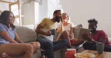 Çok ırklı bir arkadaş grubu hafta sonları evde oturup, kanepeye oturup, abur cubur yiyor, konuşuyor ve gülümsüyor ve dijital tablet kullanıyor.