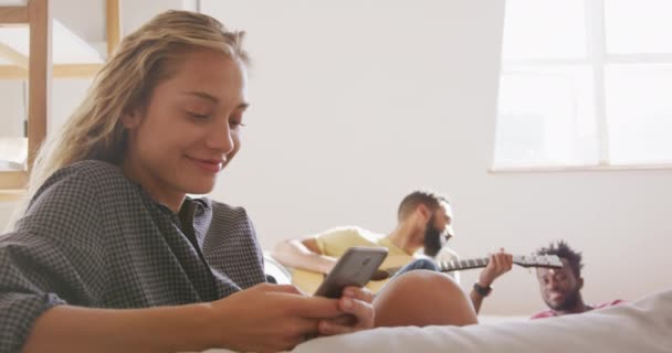 白种人女人周末在家里休息 坐在沙发上 用智能手机和朋友们一起在后台弹吉他 — 图库视频影像