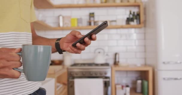 混血男子在家里休息 站在厨房里 坐在桌旁 拿着一杯茶 用智能手机 隔离隔离中的社会疏离 — 图库视频影像