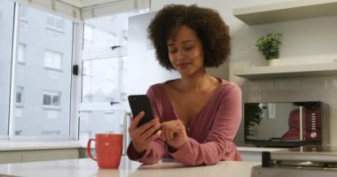 Karışık ırktan bir kadının evde vakit geçirmesinin düşük açılı görüntüsü. Mutfakta bir masada oturup kahve içmesi ve akıllı telefon kullanması, gülümsemesi.