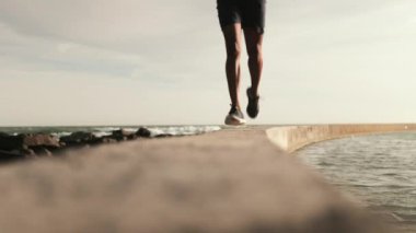 Afrika kökenli Amerikalı bir adam spor kıyafetleri giyiyor, güneşli bir günde kumsalda egzersiz yapıyor, deniz duvarında koşuyor, ayakkabı bağcıklarını bağlamak için duruyor.