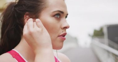 Uzun siyah saçlı, spor kıyafetleri giyen, spor yapmadan önce kulağına kulaklık takan, ağır çekimde kıvrımlı beyaz kadın..