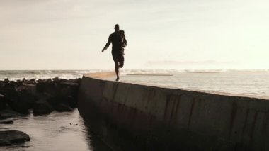 Afrika kökenli Amerikalı bir adam spor kıyafetleri giyiyor, kumsalda egzersiz yapmaktan hoşlanıyor, deniz duvarında koşuyor, arka planda, yavaş çekimde.