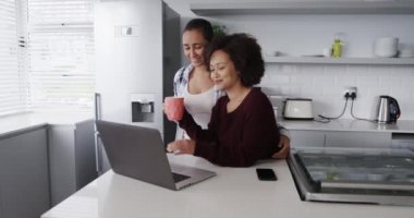 Karışık ırktan kadın bir çiftin kahve içerken ve mutfakta dizüstü bilgisayar kullanırken eğlenmesini izlemek.