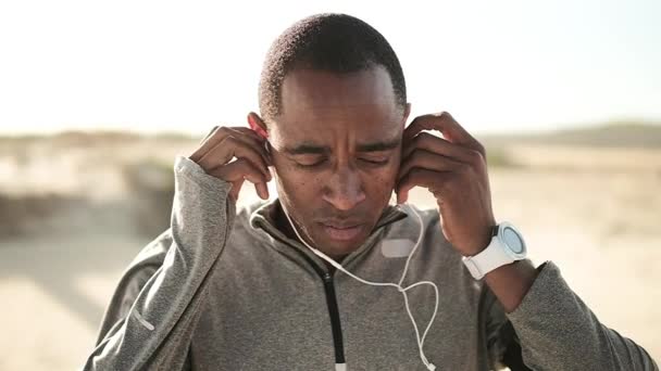 身穿运动服的非裔美国人 在阳光灿烂的海滩上很早就开始锻炼 戴上耳机 面带微笑 — 图库视频影像