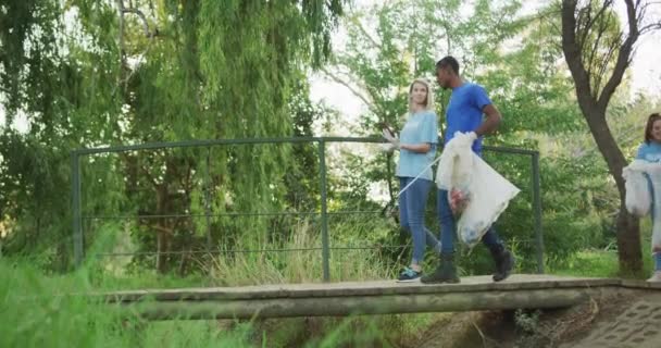 在一个阳光明媚的夜晚 一群多种族的环保志愿者在一座桥上行走 清理着一条河 农村环境中的生态和社会责任 — 图库视频影像