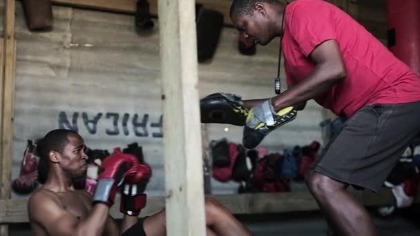 町のボクシングジムでボクシングの手袋の訓練を身に着けているアフリカ系アメリカ人男性ボクサー座ってパンチパッドを身に着けているアフリカ系アメリカ人男性トレーナーの手でパンチを投げる スローモーション — ストック動画