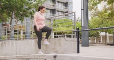 Uzun siyah saçlı kıvrımlı beyaz kadın spor kıyafetleri giyiyor, şehirde egzersiz yapıyor, bağcıklarını bağlıyor, bacağını duvara dayıyor, ağır çekimde..