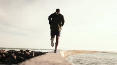 Afrika kökenli Amerikalı bir adamın spor kıyafetlerini giymesini kumsalda egzersiz yapmasını, deniz duvarında koşmasını, ağır çekimde koşmasını.