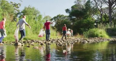 Kırsal kesimde güneşli bir günde nehri temizleyip çöp toplayan çok ırklı bir koruma gönüllüsü grubunun arka görüntüsü. Kırsal bir ortamda ekoloji ve sosyal sorumluluk.