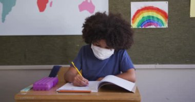 Okulda maskesini takmış, kitap yazan melez bir kız. Covid19 Coronavirus salgını sırasında temel eğitim sosyal güvenlikten uzaklaşma yavaş çekimde.