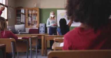 Sınıfta çok ırklı çocuklar el kaldırıyor, maske takıyor. Covid19 Coronavirus salgını sırasında temel eğitim sosyal güvenlikten uzaklaşma yavaş çekimde.