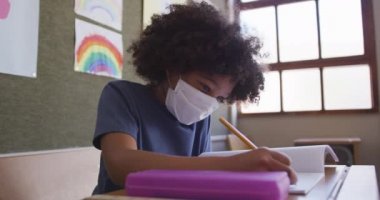 Okulda maskesini takmış, kitap yazan melez bir kız. Covid19 Coronavirus salgını sırasında temel eğitim sosyal güvenlikten uzaklaşma yavaş çekimde.