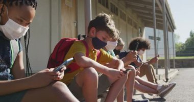 Çok ırklı bir grup çocuk akıllı telefonlarını kullanıyor, okulda yüz maskeleri takıyorlar. Covid19 Coronavirus salgını sırasında temel eğitim sosyal güvenlikten uzaklaşma yavaş çekimde.