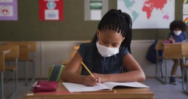 Okulda maskesini takmış, kitap yazan melez bir kız. Covid19 Coronavirus salgını sırasında temel eğitim sosyal güvenlikten uzaklaşma yavaş çekimde.