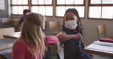 Yüz maskesi takan iki kız okulda dirseklerine dokunarak birbirlerini selamlıyorlar. Covid19 Coronavirus salgını sırasında temel eğitim sosyal güvenlikten uzaklaşma yavaş çekimde.