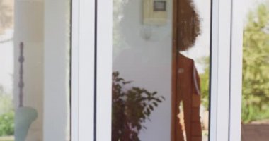 Afrika kökenli Amerikalı son sınıf öğrencisi, yüz maskesi takmış, pencerenin arkasından ailesini selamlıyor. Coronavirus Covid 19 karantina sürecinde sosyal uzaklık ve kendini izole etme.