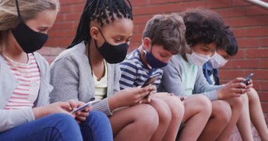 Mola sırasında duvarda oturan akıllı telefonları kullanarak yüz maskesi takan çok ırklı çocuklardan oluşan bir grup. Covid19 Coronavirus salgını sırasında temel eğitim sosyal güvenlikten uzaklaşma yavaş çekimde