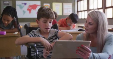 Kafkasyalı engelli çocuk tekerlekli sandalyesinde oturuyor, öğretmen ona okulda tablet gösteriyor. Covid19 Coronavirus salgını sırasında temel eğitim sosyal güvenlikten uzaklaşma yavaş çekimde.